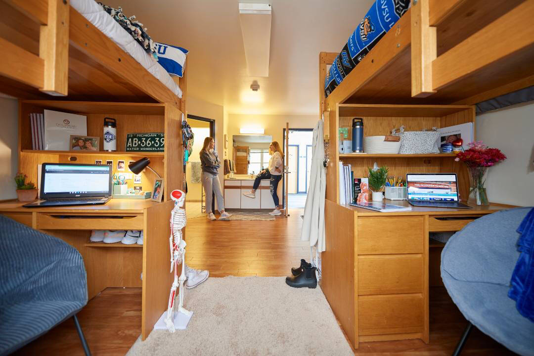 Roommates laugh together in Suite Style unit, with two lofted beds, desks, sinks, and a bathroom in the background.